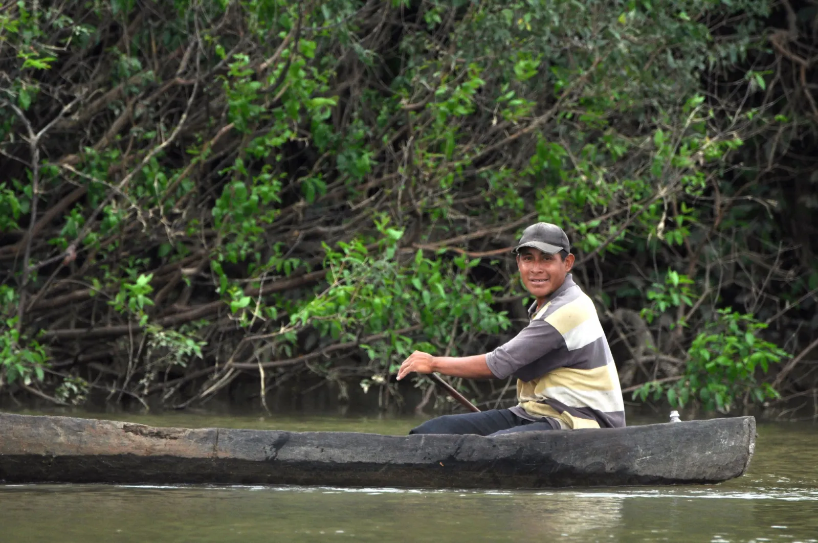 A person paddling a wooden canoe beside a wooded riverbank